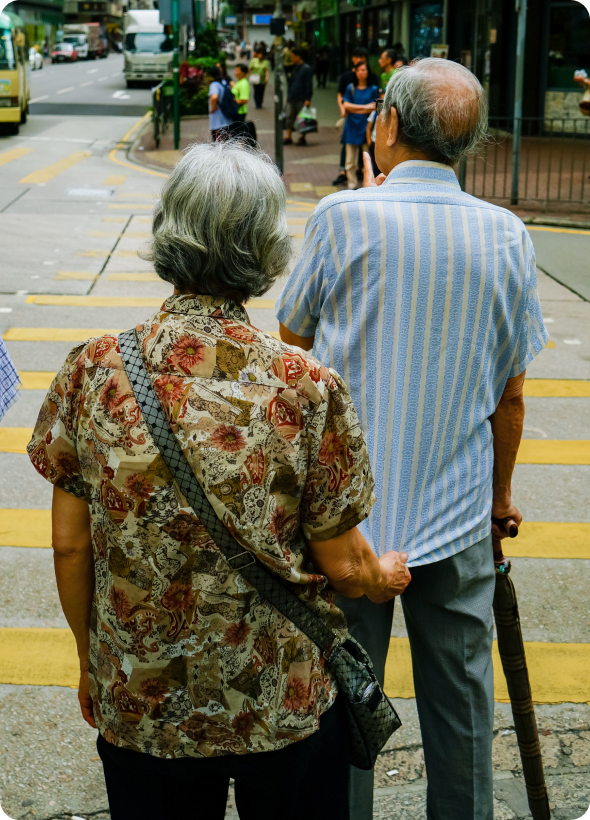 An elderly couple waiting to cross the street and the woman stands behind man pulling him back