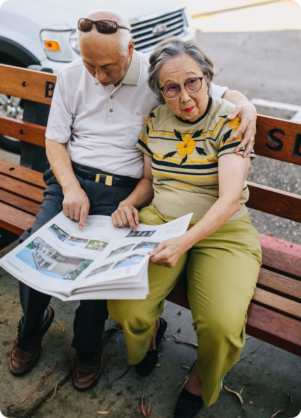 An elderly couple sits together on a bench reading a newspaper, the man's arm draped across the woman's shoulders