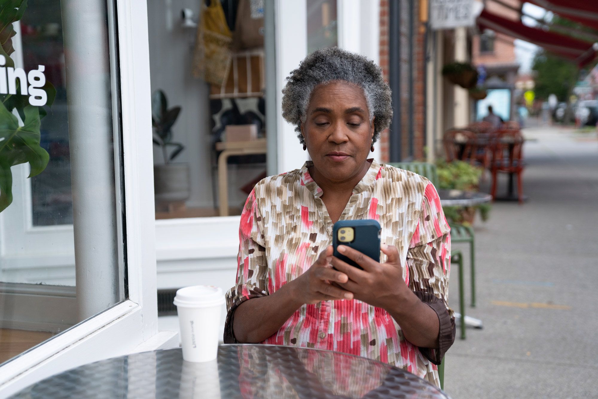 A woman sits at a table with a coffee while looking at her phone