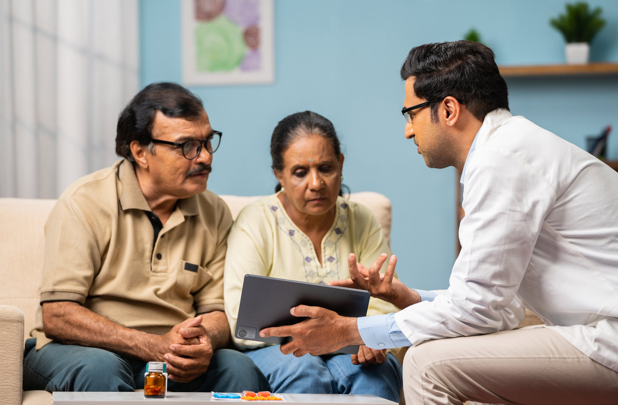 Indian doctor explaining from digital tablet to senior couple about medical report at home – concept of technology, health care support and home consultation.